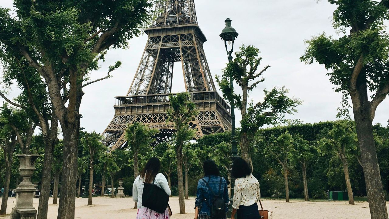 Three people stand in a park facing the Eiffel Tower, amid trimmed trees under a cloudy sky.