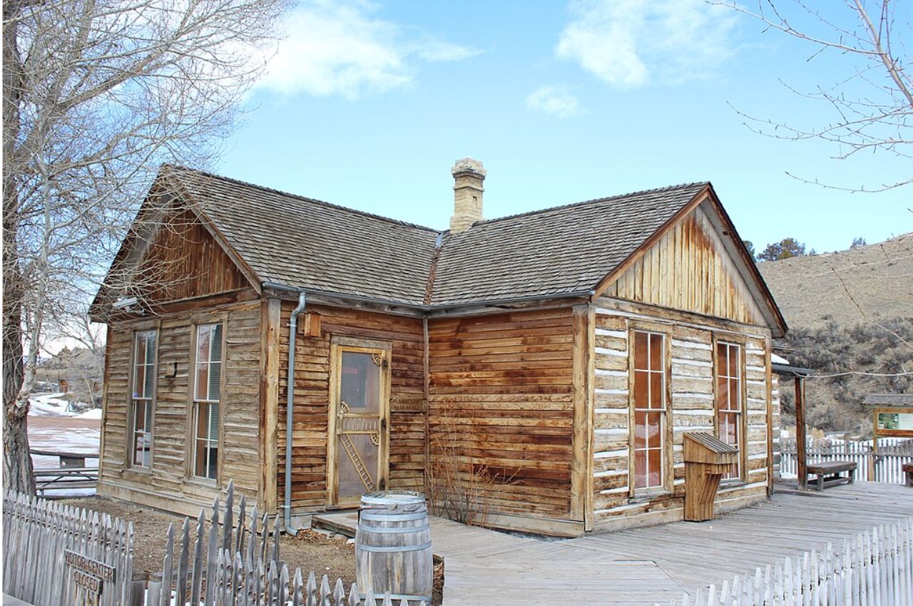 A small, weathered wooden house with a shingled roof and chimney stands inside a picket fence in a rural area under a blue sky.