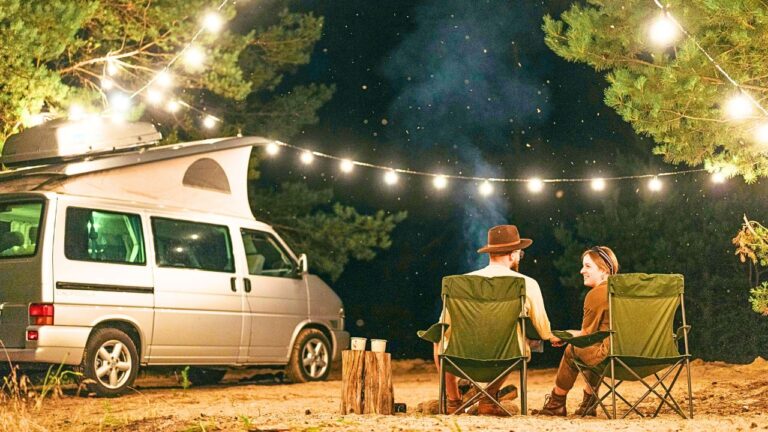 Two people sit in camping chairs by a van under string lights at night, surrounded by trees, mugs, and a log.