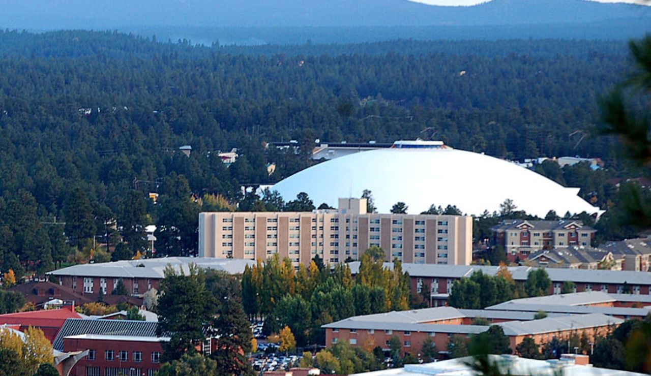 Aerial view of the Walkup Skydome at Northern Arizona University in Flagstaff, surrounded by campus buildings, pine forest, and distant mountain ranges.