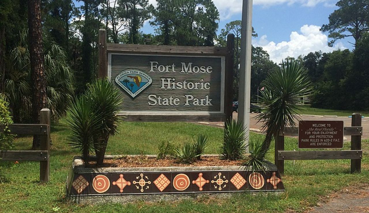 Entrance sign for Fort Mose Historic State Park in Florida, surrounded by palm plants and trees.