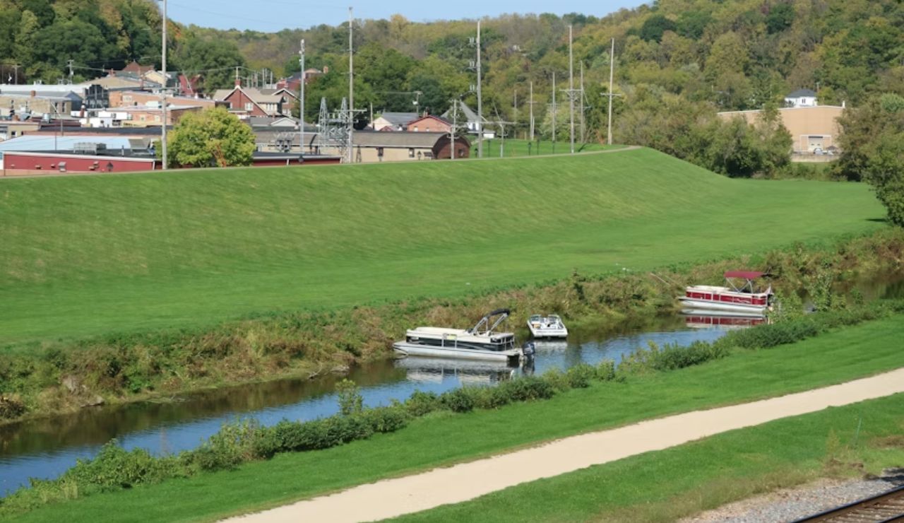 Small boats docked on a calm river beside a green levee with a town and wooded hills in the background.