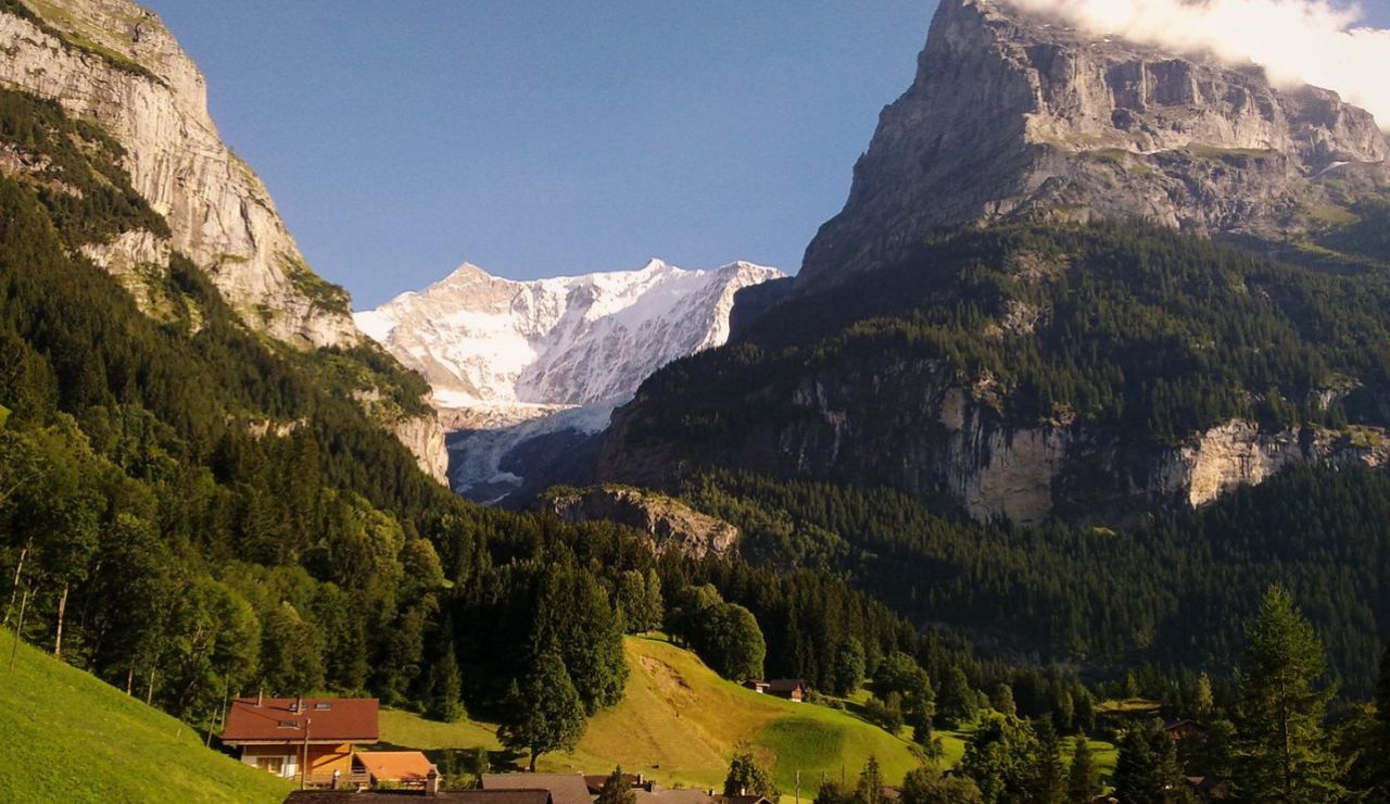 A peaceful mountain village nestled in lush green hills, with towering cliffs and snow-capped peaks rising in the distance.