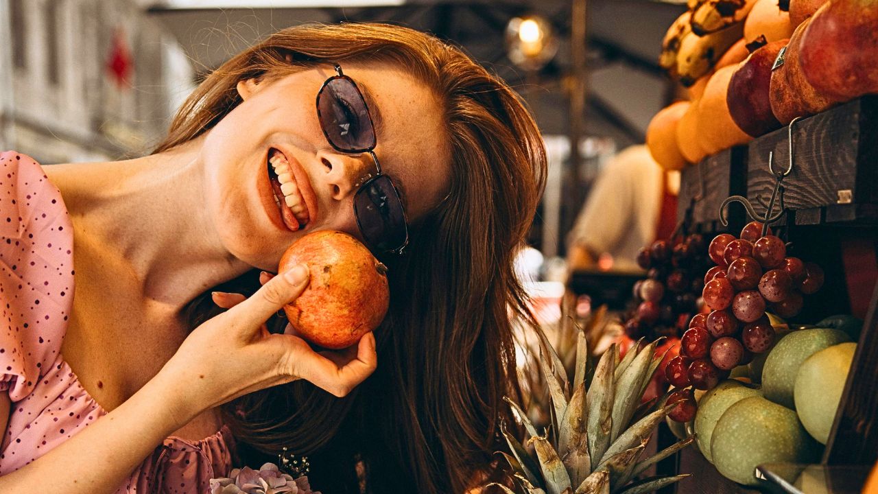 Wearing sunglasses and a pink dress, a person smiles while holding a pomegranate to their face at a fruit market surrounded by various fruits.