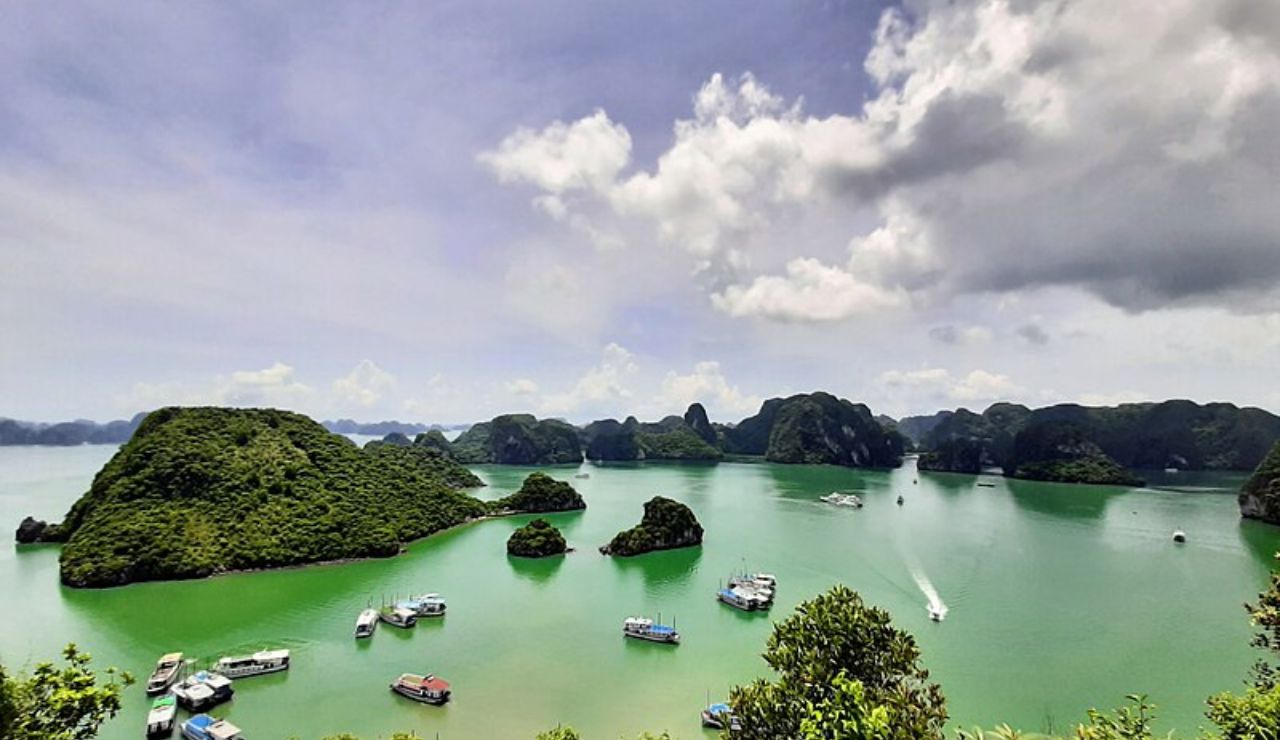 Boats cruise through the emerald waters and limestone islands of Ha Long Bay, Vietnam, under a partly cloudy sky.