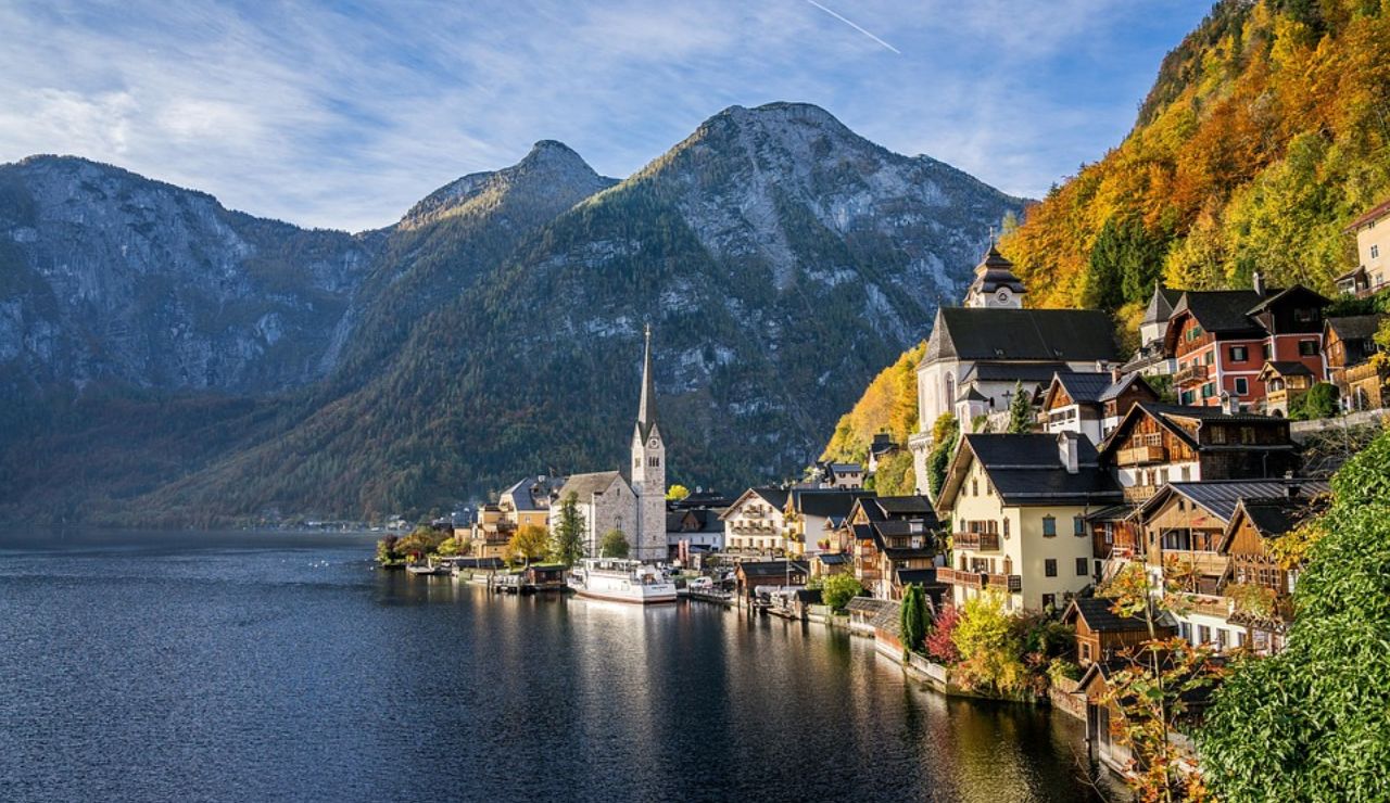 Picturesque view of Hallstatt village in Austria with traditional alpine houses, a church steeple, and autumn-colored mountains reflecting in the lake.