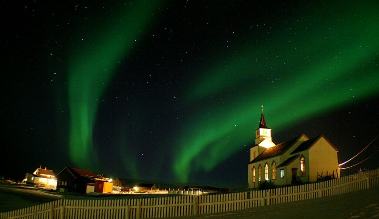 Northern lights glowing above a church and snow-covered village in Hillesøy, Norway.
