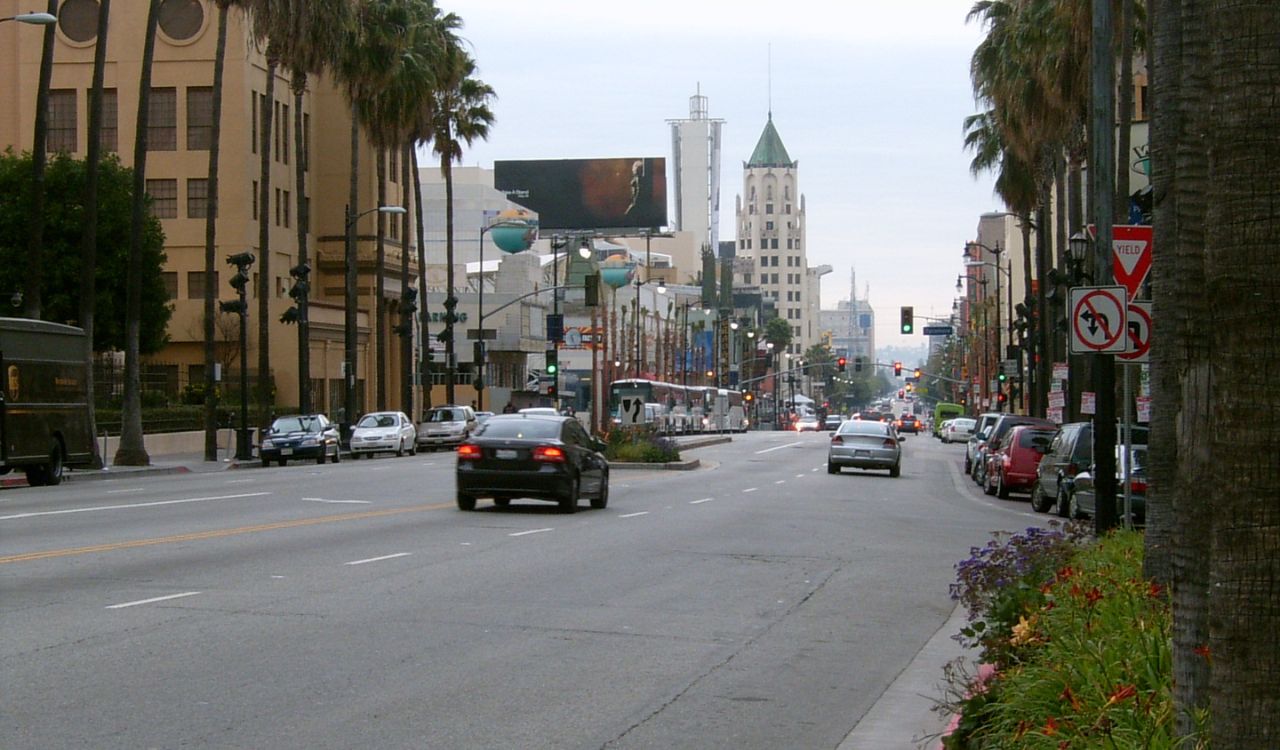 Palm-lined street with traffic heading toward a city skyline under cloudy skies.