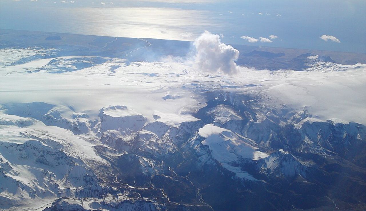 Aerial view of Eyjafjallajökull volcano in Iceland, partially covered by a glacier, with steam rising from its crater and the North Atlantic Ocean shimmering in the distance.