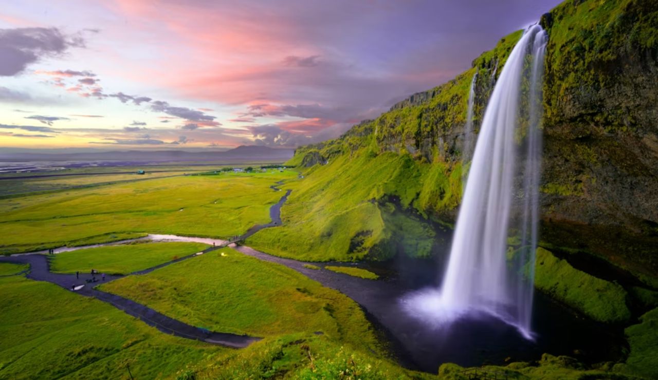 Seljalandsfoss waterfall cascading over a mossy cliff at sunset in southern Iceland, with a vibrant sky and lush green landscape below.