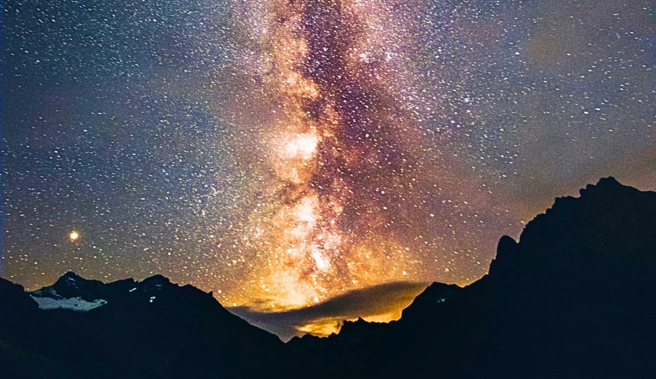 The colorful Milky Way arches across a starry night sky above dark mountain silhouettes.
