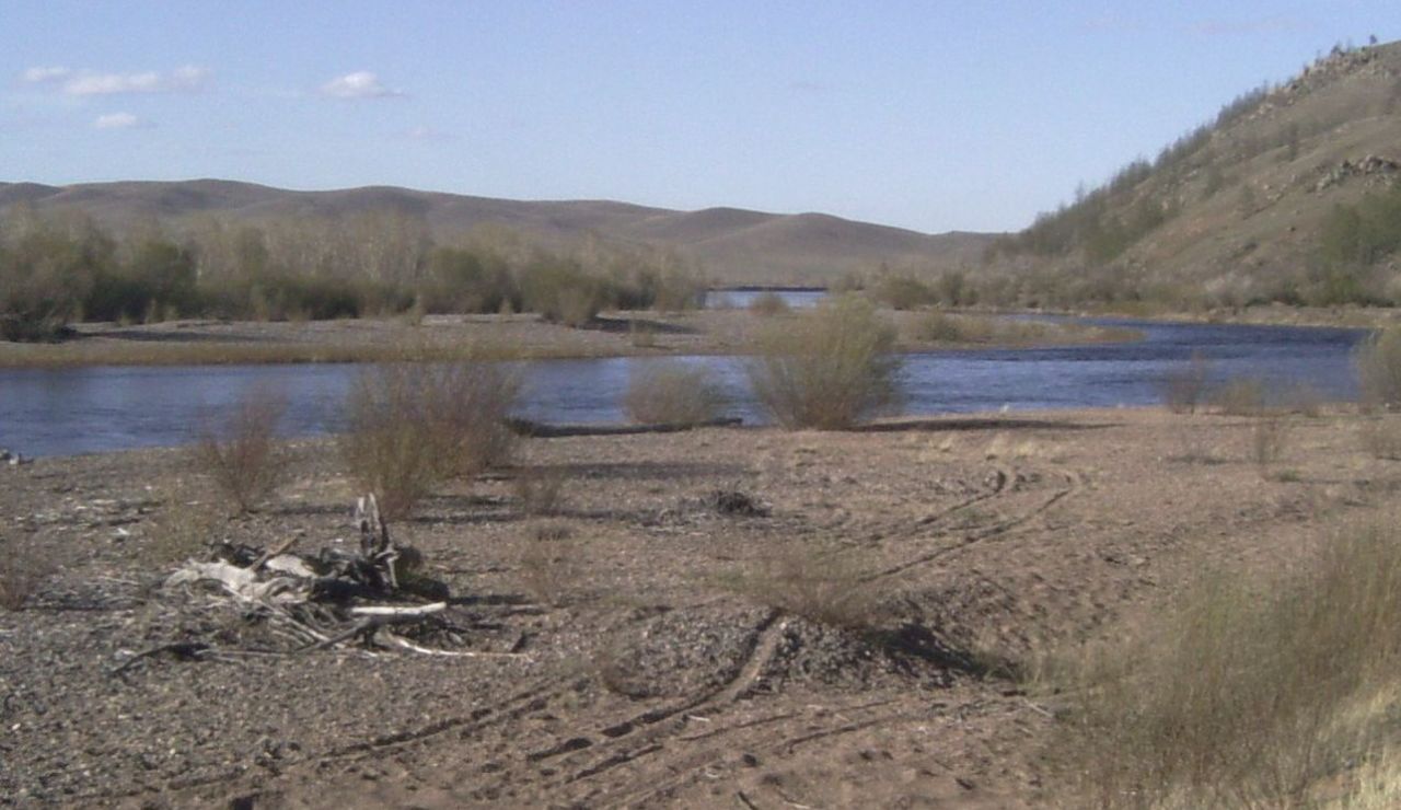 A calm river winds through a dry, rocky landscape with scattered shrubs and distant rolling hills under a clear blue sky.