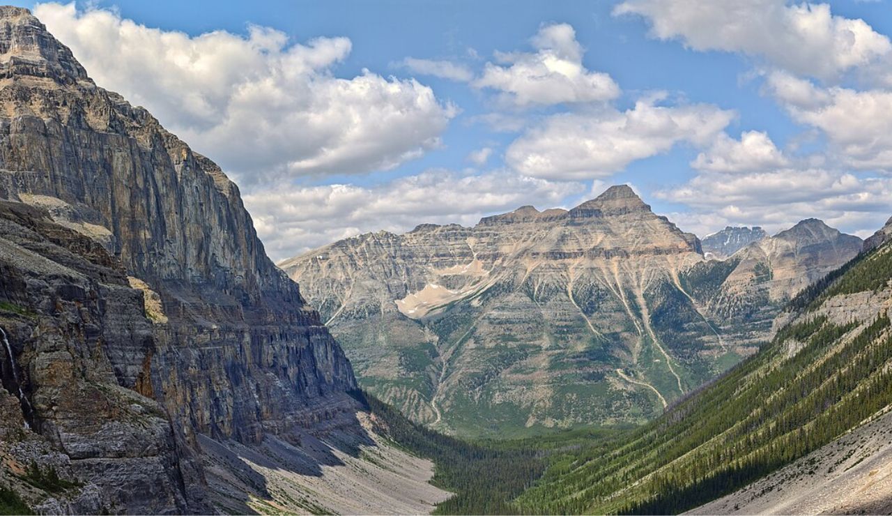 Rugged mountain peaks and deep valleys in Kootenay National Park, British Columbia, with cascading waterfalls and forested slopes under a partly cloudy sky.