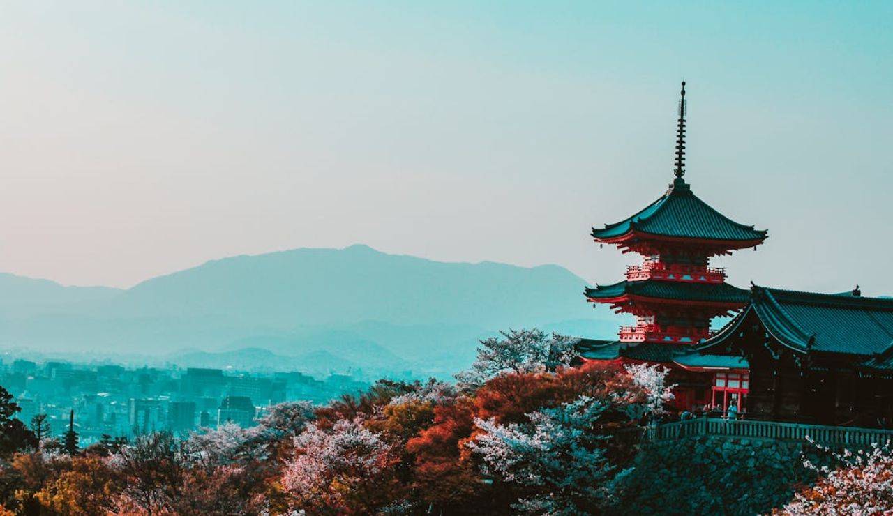 Traditional red Japanese pagoda surrounded by cherry blossoms in full bloom overlooking Kyoto city with misty mountains in the background.