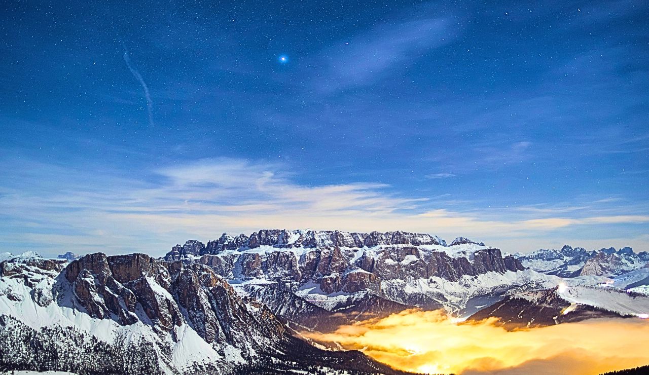 Snow-covered mountains rise beneath a starry sky, with a bright star overhead and glowing clouds illuminating the valley below.