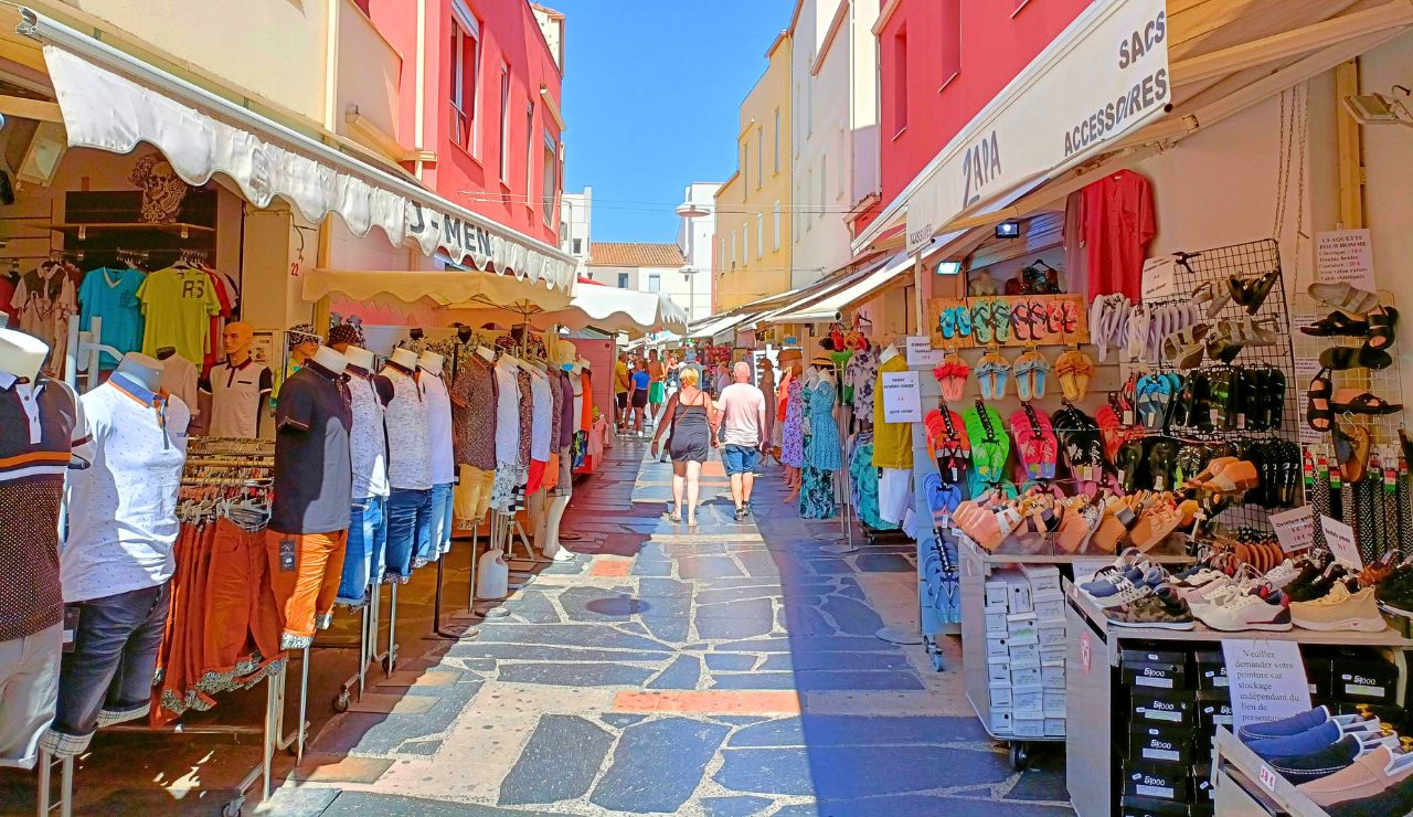 A bustling outdoor marketplace features colorful buildings, clothing and shoe displays lining a stone-paved street filled with pedestrians in bright sunlight.