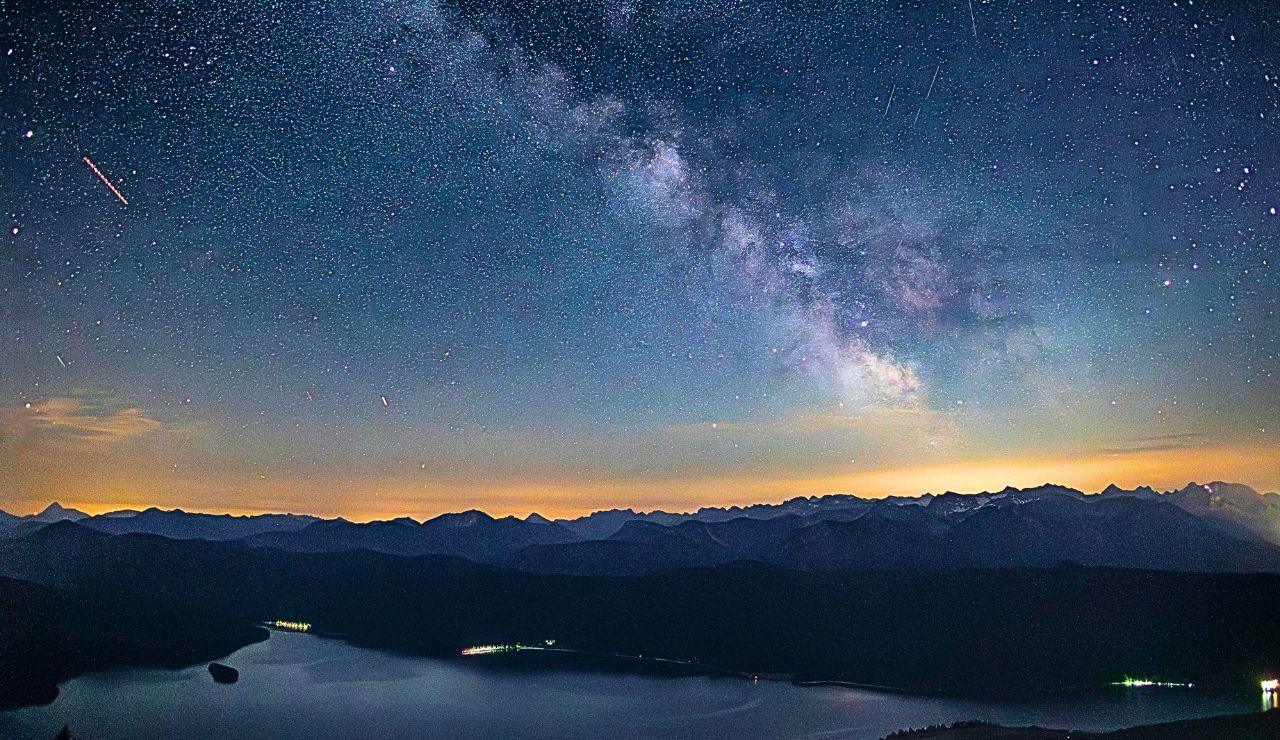 The Milky Way shines above silhouetted mountains and a calm lake, with distant shoreline lights.