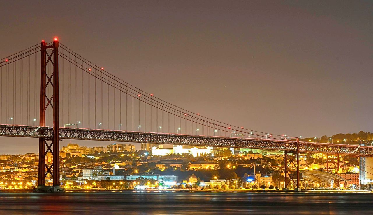 A brightly lit suspension bridge spans a river at night, with a cityscape glowing in the background.