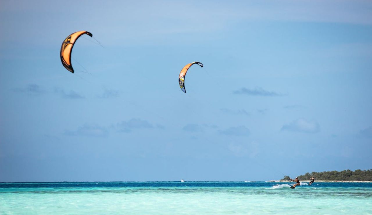 Two people kite surfing over clear turquoise waters in Los Roques, Venezuela, with a tropical shoreline and bright blue sky in the background.