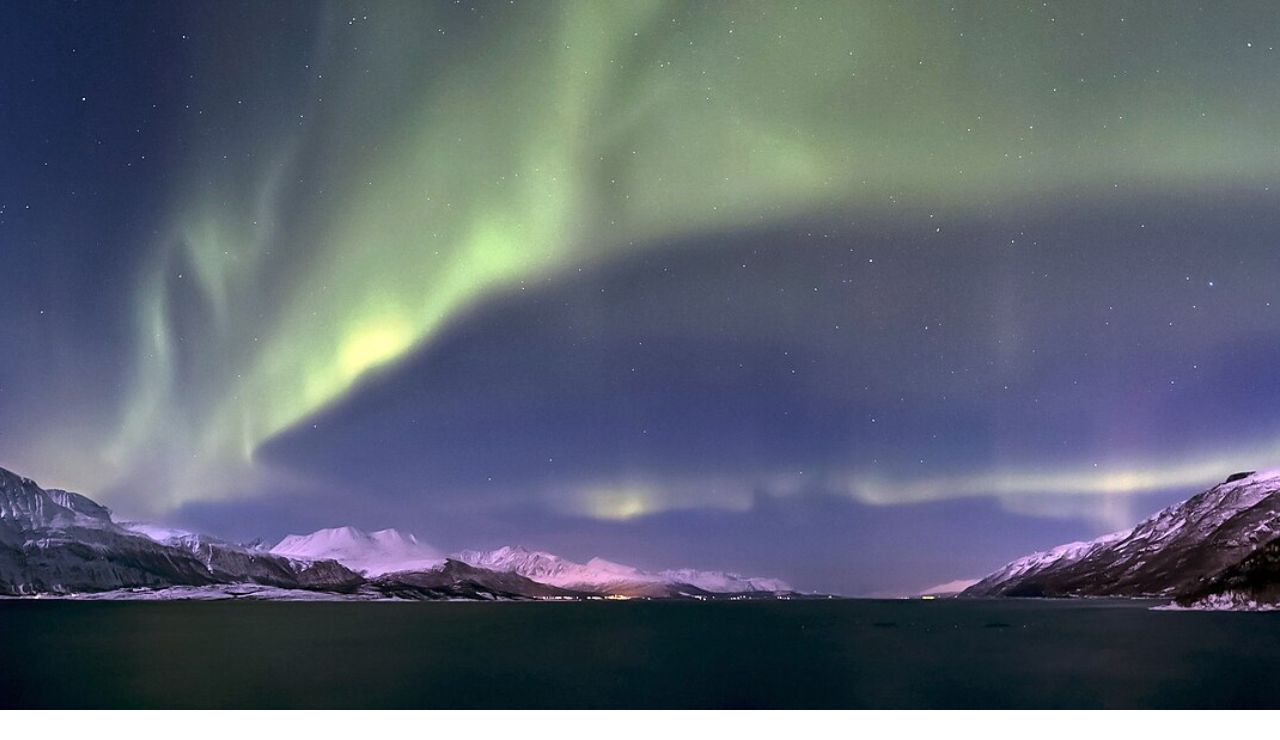 Northern lights glowing above snow-covered peaks and fjord waters in the Lyngen Alps, Norway.