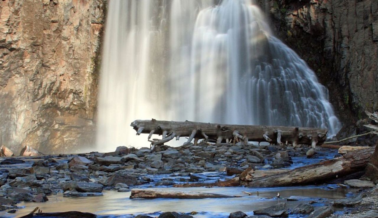 A powerful waterfall cascades over a rocky cliff, landing beside a large fallen log and scattered stones in a rugged, natural pool.