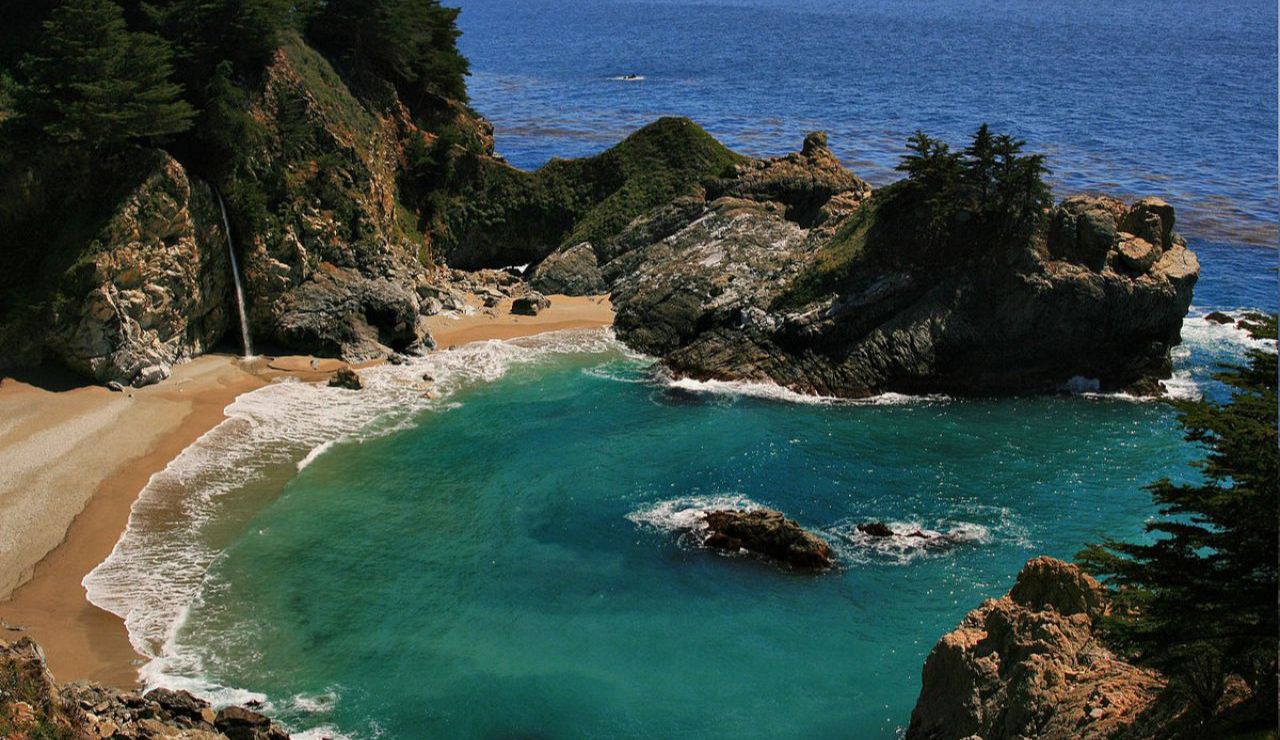 A serene cove with turquoise waters, a sandy beach, and a waterfall flowing directly onto the shore—surrounded by rugged cliffs and lush greenery.