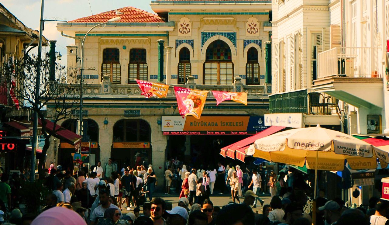 A crowded street unfolds before the Büyükada ferry terminal in Istanbul, with people walking amid colorful banners and historic buildings.