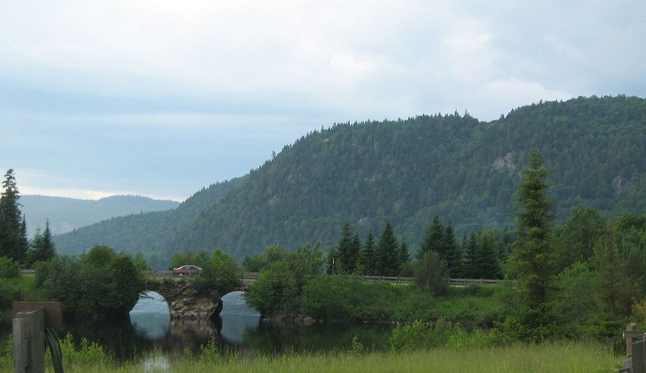 Stone arch bridge over a calm river surrounded by forested hills in Parc national des Hautes-Gorges-de-la-Rivière-Malbaie, Quebec, Canada.