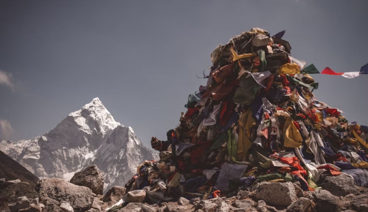 Colorful prayer flags drape a stone cairn in the Himalayas with a snow-covered peak in the background.