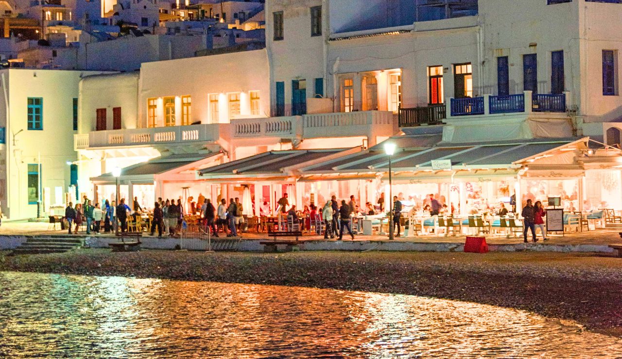 People dine at outdoor waterfront restaurants at night, with lights from white buildings reflecting on the water.