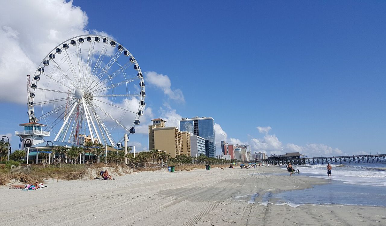 Beachfront with a giant Ferris wheel, high-rise hotels, and people relaxing by the ocean under clear blue skies.
