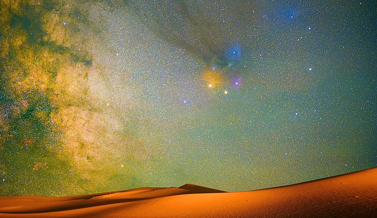A starry night sky with colorful nebulae glows above softly lit sand dunes.