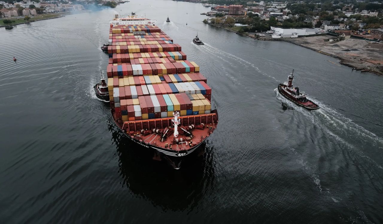 A massive cargo ship, stacked high with colorful containers, is escorted through a waterway by tugboats near the coast.