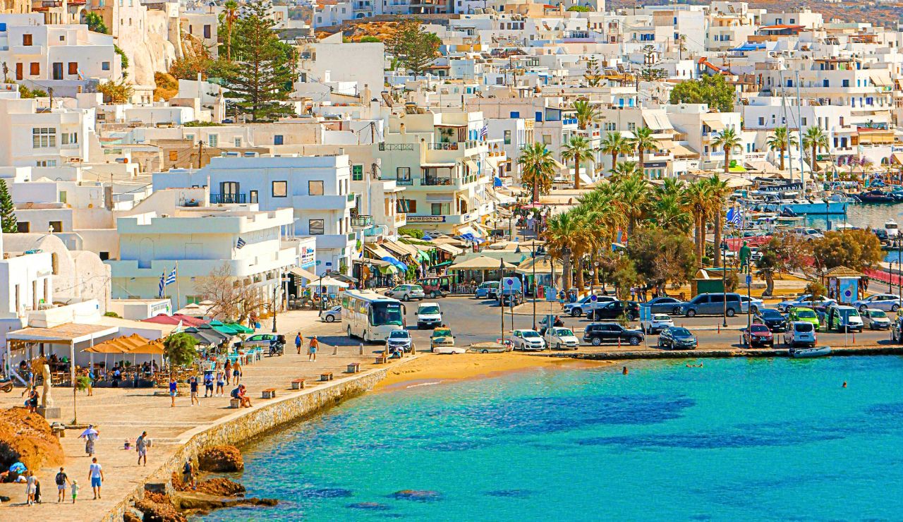 Coastal town with white buildings, a lively promenade lined with palm trees, parked cars, people walking, and a marina by turquoise water.