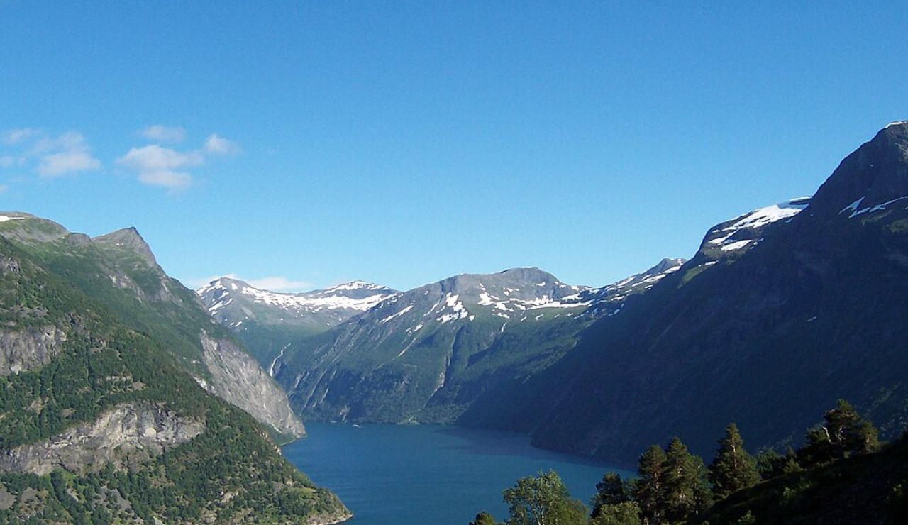 Aerial view of Geirangerfjord in Norway, showcasing deep blue waters, steep cliffs, snow-capped peaks, and lush green forests under a clear blue sky.