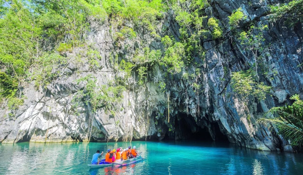 Tourists in a boat entering the turquoise waters of Puerto Princesa Underground River in Palawan, Philippines, surrounded by limestone cliffs and lush jungle.
