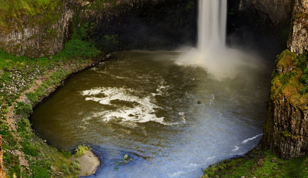 A tall, narrow waterfall drops into a wide, round basin surrounded by steep rocky cliffs and lush green vegetation.