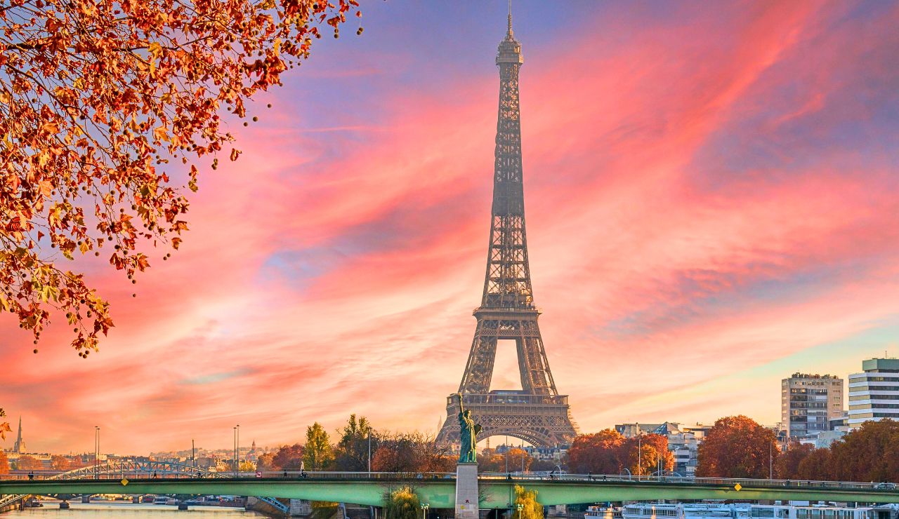 The Eiffel Tower rises in the background behind autumn trees, a bridge, and the Seine River at sunset.