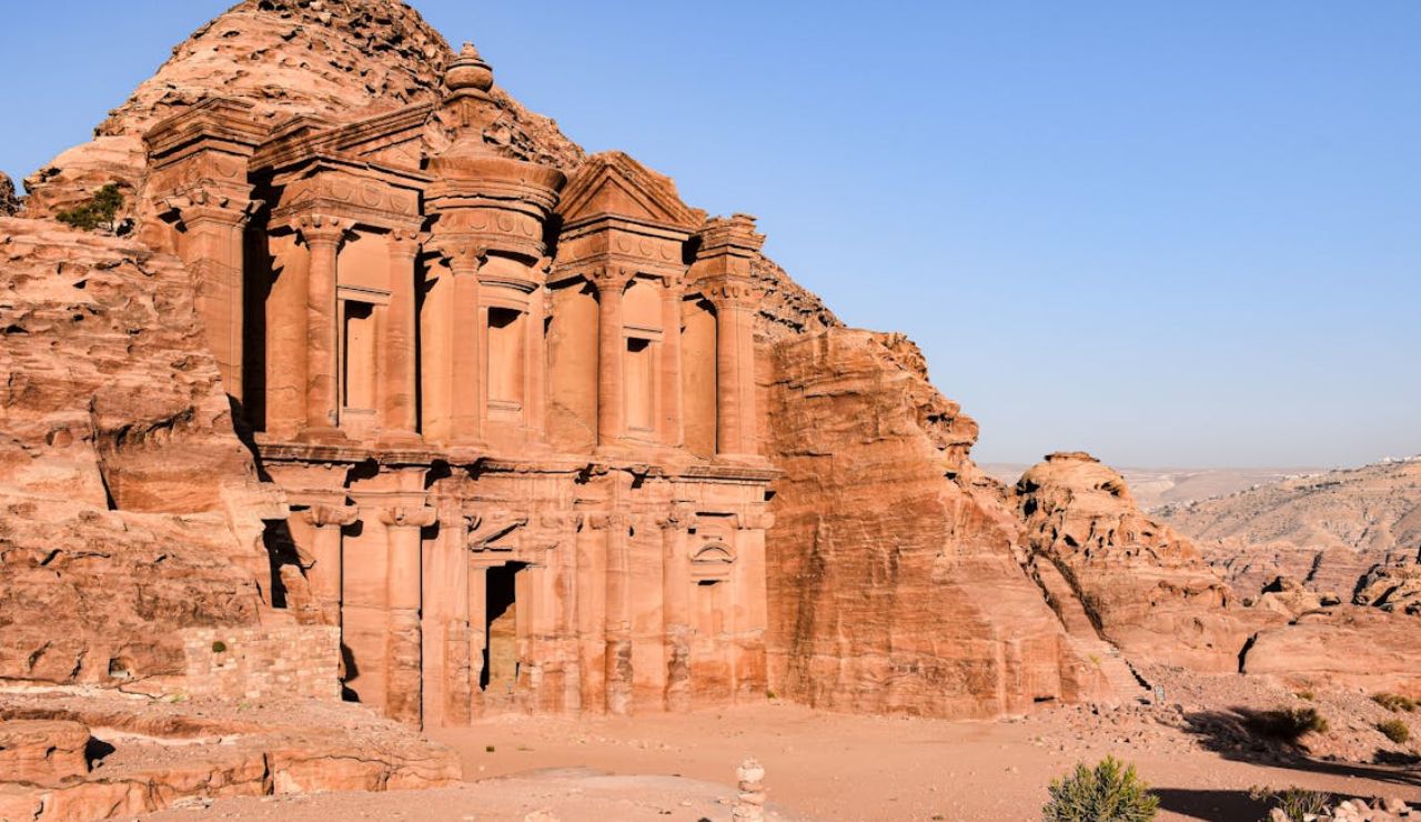 The Monastery (Ad Deir), an ancient rock-cut structure in Petra, Jordan, carved into the rose-red cliffs of the mountains under a clear blue sky.