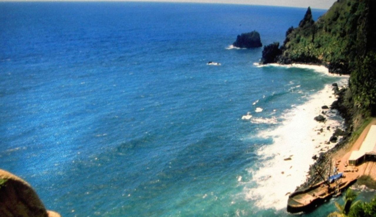 Waves crash on a rocky coast, with clear blue sea and a quiet dock below lush green cliffs.