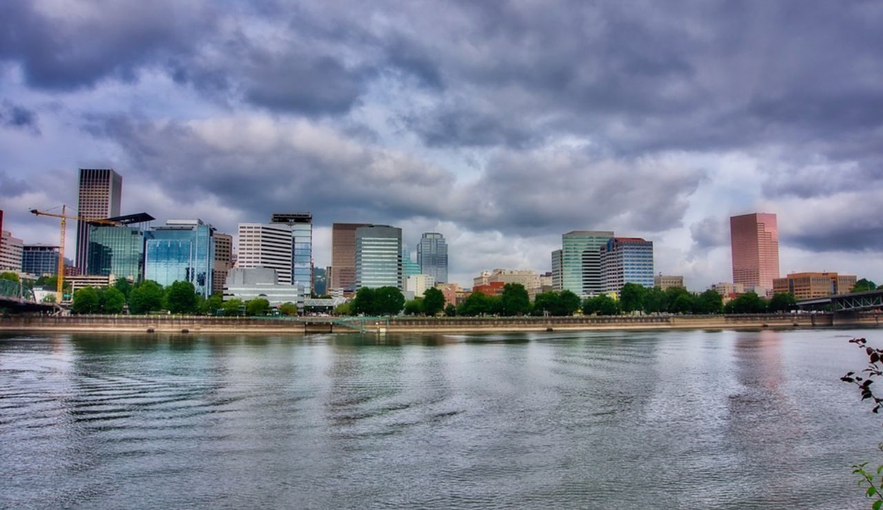 Portland, Oregon city skyline with modern buildings reflected in the Willamette River under a dramatic cloudy sky.