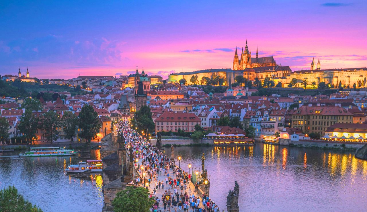 At sunset, a large crowd crosses a historic stone bridge over a river, with illuminated castle buildings in the background.