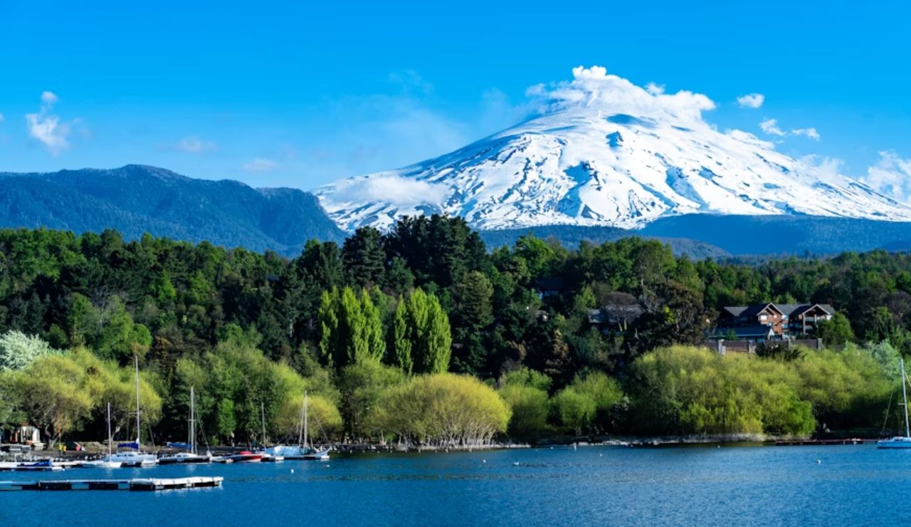 Snow-capped Villarrica Volcano rising behind lush forest and lake in Pucón, Chile, with sailboats docked along the shoreline under a clear blue sky.
Snow-capped Villarrica Volcano rising behind lush forest and lake in Pucón, Chile, with sailboats docked along the shoreline under a clear blue sky.