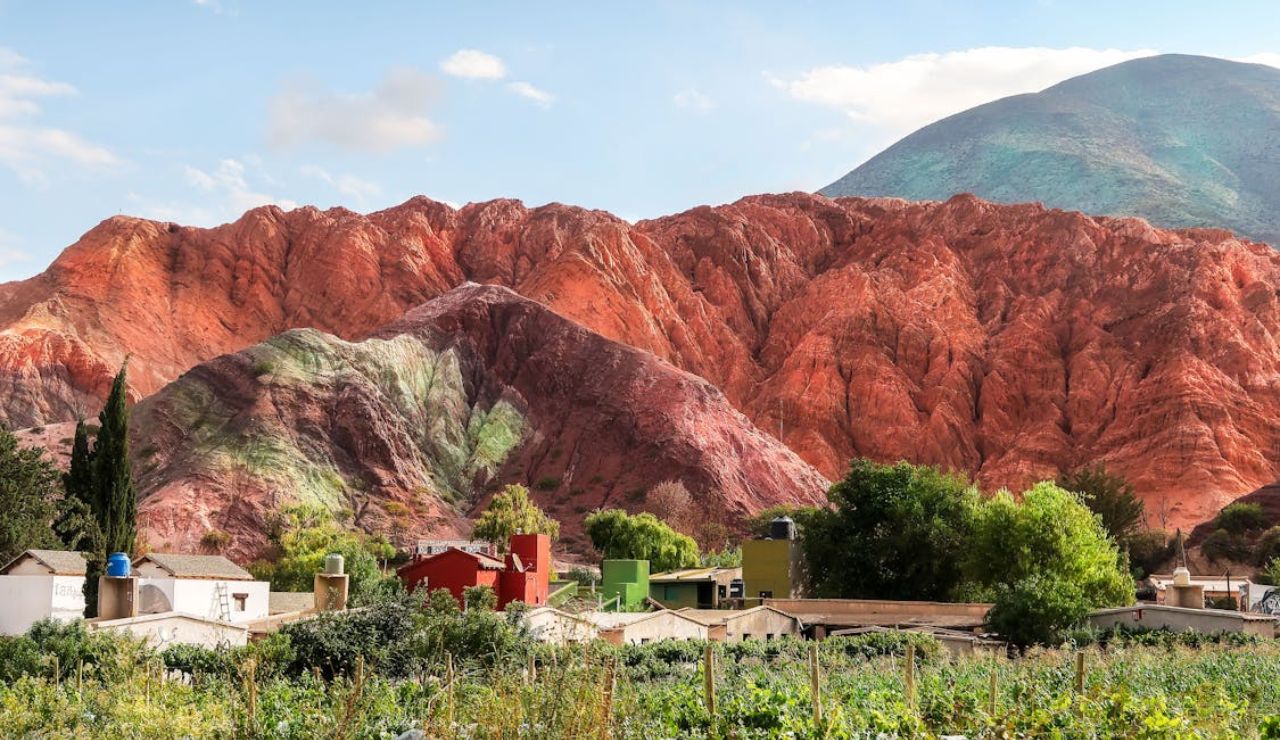 Colorful rock formations of Quebrada de Humahuaca rise behind a small village with vibrant houses and green fields in northern Argentina.