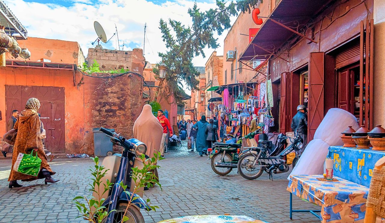 A busy street market features people walking, colorful textiles, parked motorcycles, and stalls lining orange buildings.