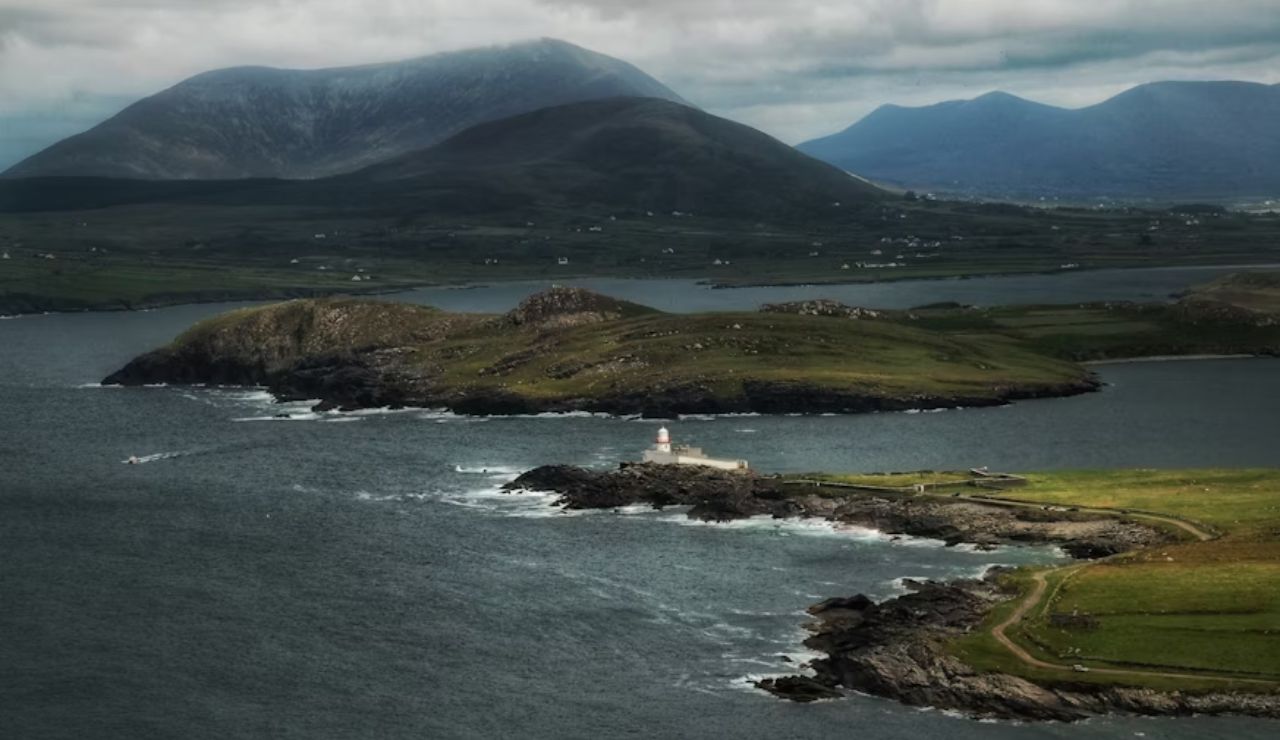 Dramatic coastal view from the Ring of Kerry in Ireland, featuring a lighthouse on a rocky peninsula, lush green hills, and distant mountains under a moody sky.