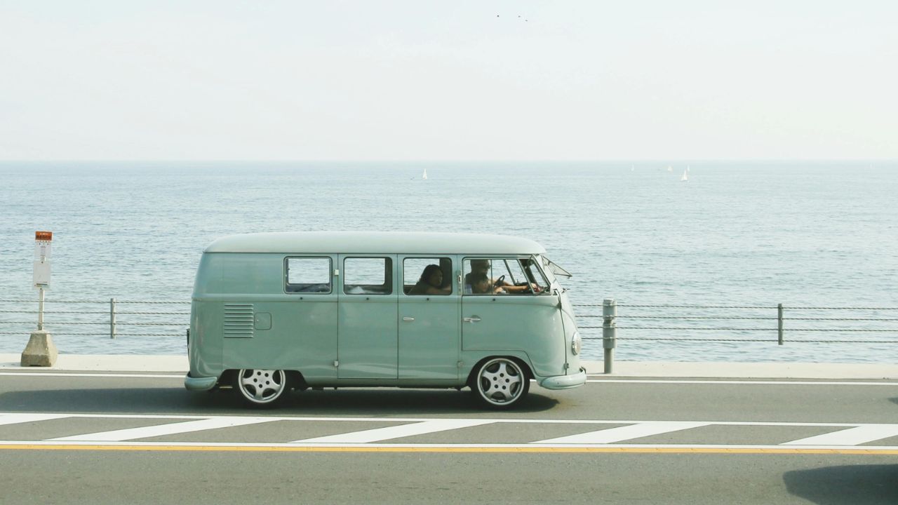 A light green vintage van drives along a coastal road with the ocean and sailboats in the background.