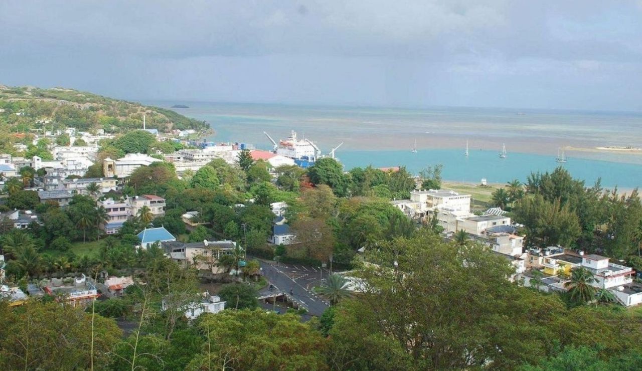 A coastal town filled with greenery and white buildings, overlooking a calm turquoise sea with ships docked and yachts anchored nearby.