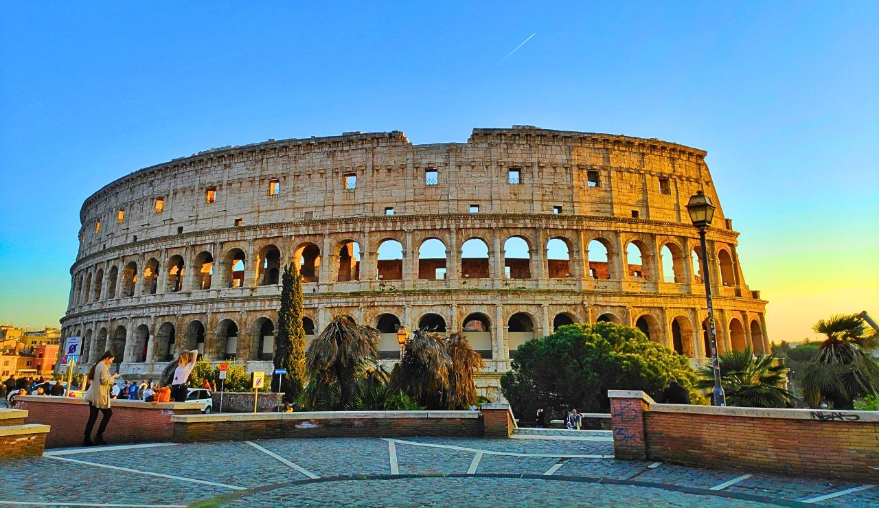 At sunset, people gather in the foreground of the Colosseum in Rome under a clear blue sky.