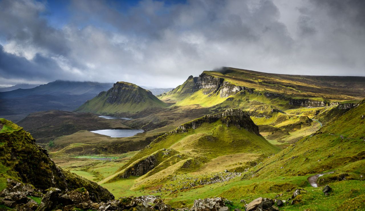 Dramatic landscape of the Quiraing landslip on the Isle of Skye, part of the Scottish Highlands, with rolling green hills, rugged cliffs, and scattered lochs under a moody sky.